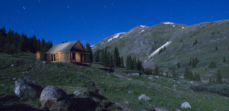 Wooden cabin in Animas Forks, Colorado at night under star trails and alpine mountain landscape