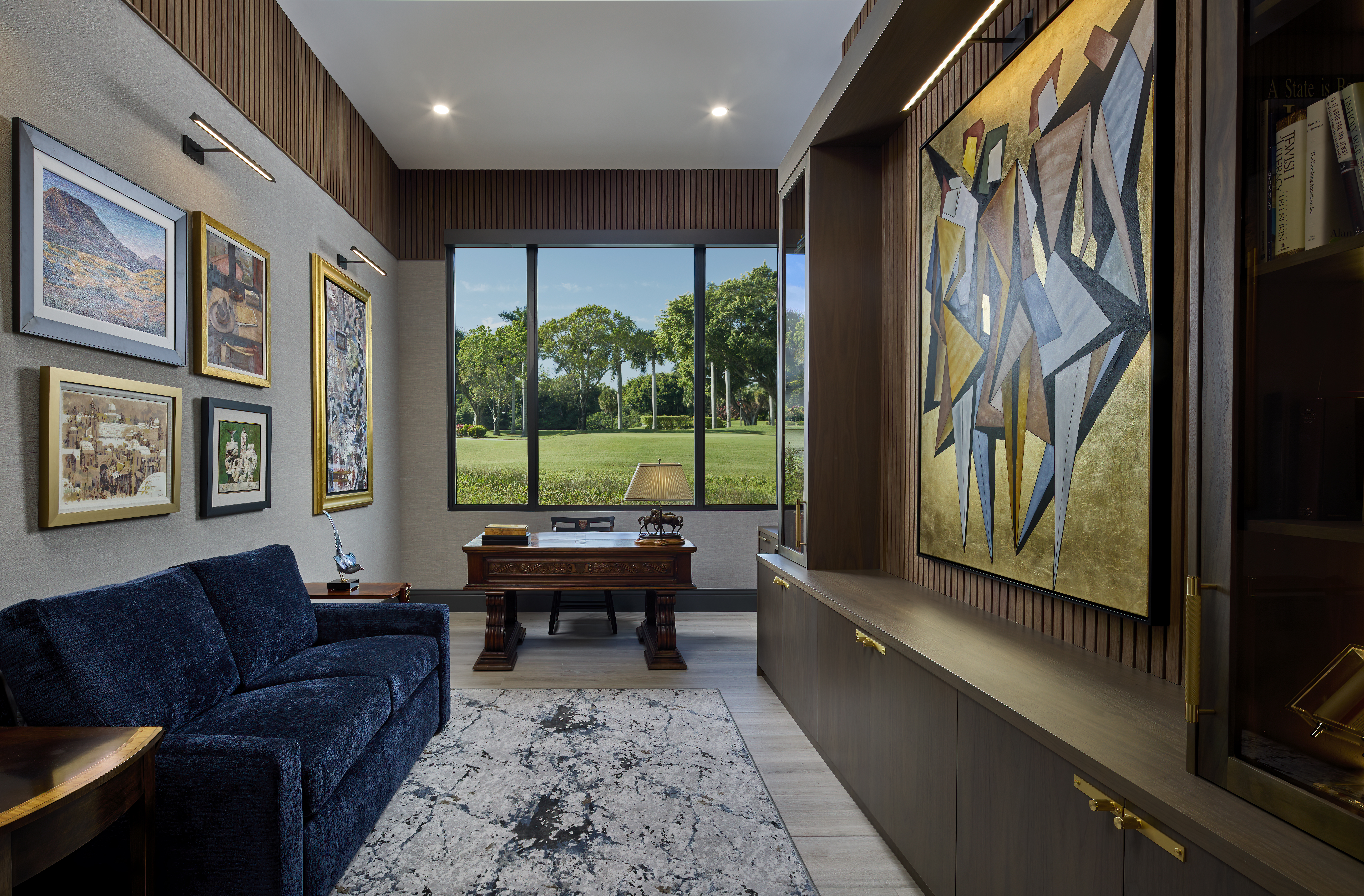 Luxury home office interior design photograph with curated art wall, built-in wood cabinetry, blue velvet sofa, large desk, and golf course view