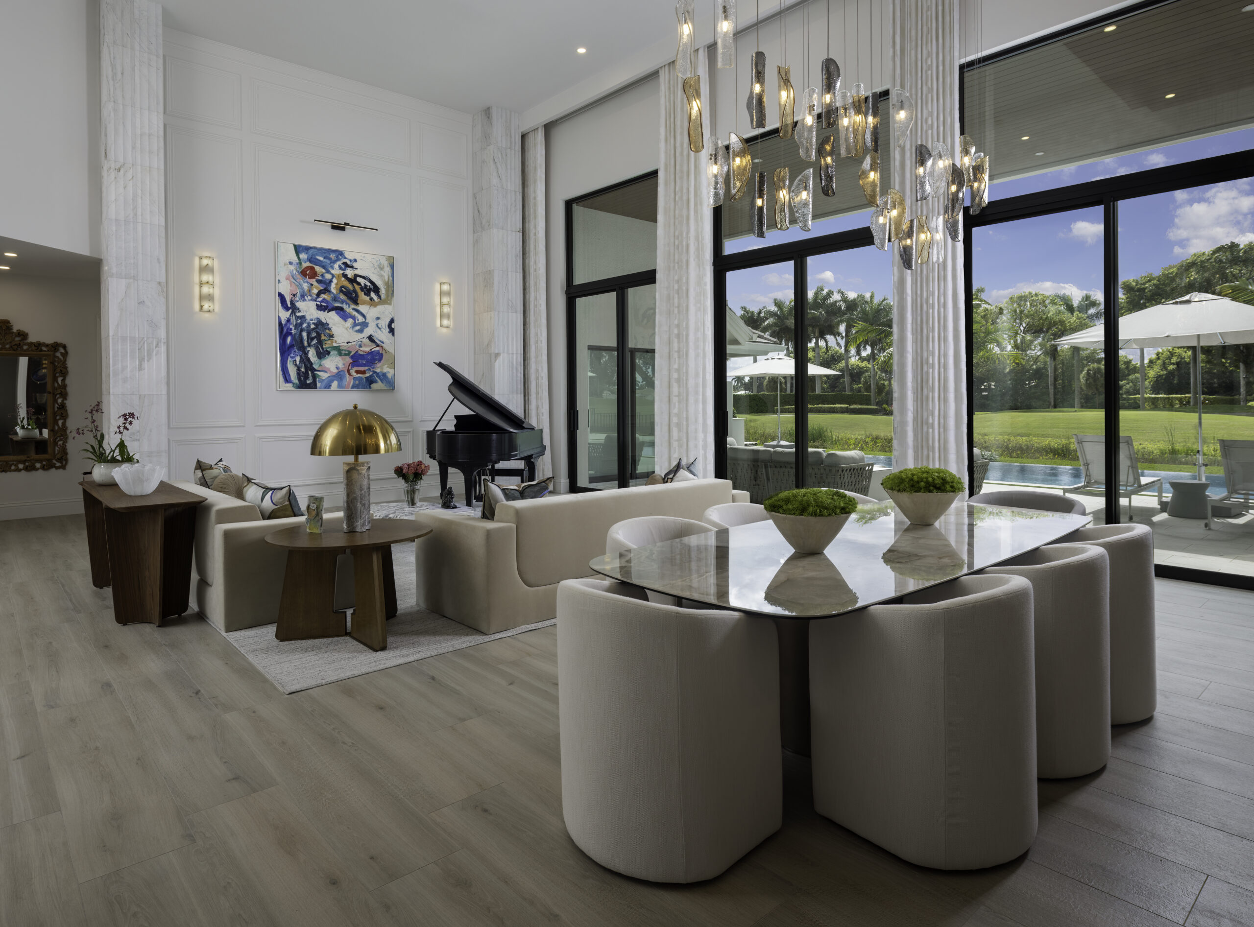 Wide luxury living room and dining room interior design photograph with grand piano, tall windows, white paneled walls, sculptural chandelier, and modern seating