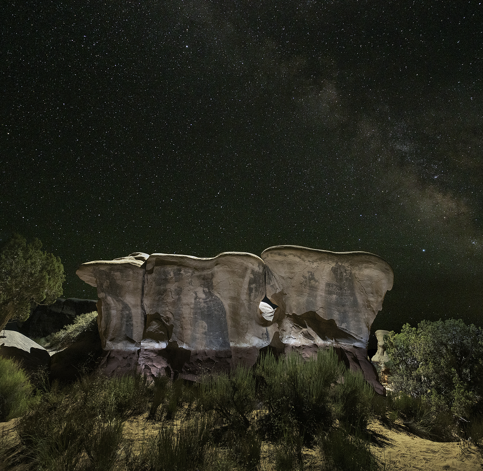 A broad sandstone formation with a window through it, lit at night beneath the Milky Way, surrounded by juniper, Devils Garden.